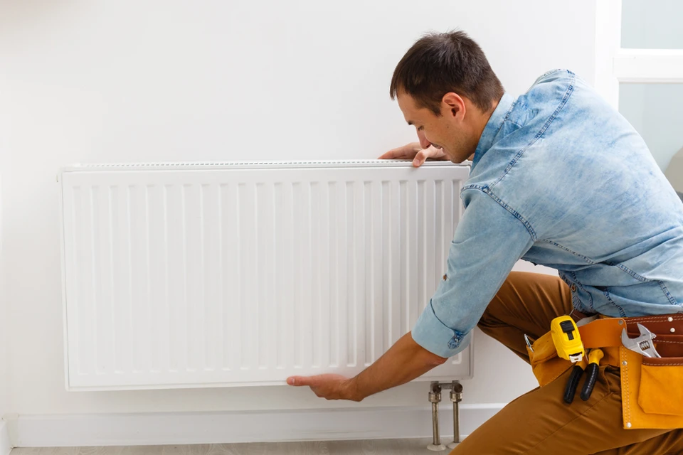 Un technicien en salopette beige et chemise en jean installe ou répare un radiateur à panneaux blanc dans un intérieur résidentiel moderne